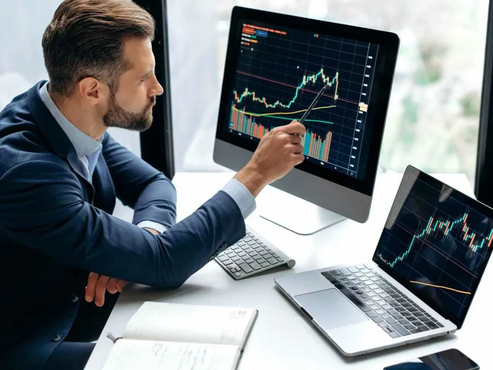 Trader pointing at a stock chart on a desktop monitor next to a laptop